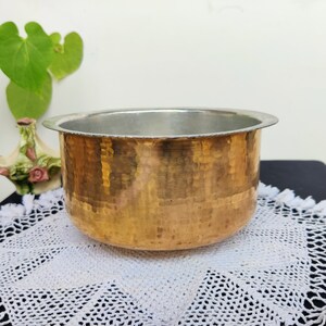 May include: A hammered copper bowl with a silver-colored interior. The bowl has a rounded shape and a wide rim. The copper exterior has a textured, dimpled surface. The bowl is sitting on a white lace doily.