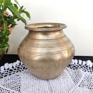 May include: A vintage brass water pot with intricate engraved designs, including elephants, on a white crocheted doily.