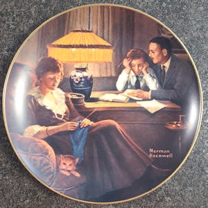 May include: Decorative plate featuring a family scene by Norman Rockwell. The image depicts a woman knitting, a child studying, and a man assisting. The plate has a gold rim and showcases warm colors, including brown, blue, and yellow.
