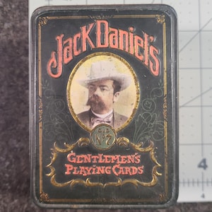 May include: A rectangular black metal tin for Jack Daniel's playing cards, featuring a portrait of a man in a hat, the text Jack Daniel's, Old No. 7, and Gentlemen's Playing Cards, positioned next to a ruler showing a height of 4 inches.
