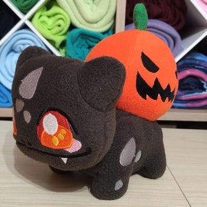 May include: A plush toy of a black and grey Pokemon character with a large orange pumpkin head with a scary face. The pumpkin head has a green stem on top.