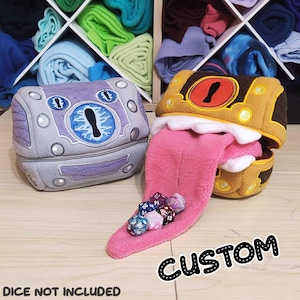 May include: Two plush dice bags, one grey and purple with an eye design, the other brown and gold with a pink tongue. A collection of dice rests on the tongue of the second bag. The word "CUSTOM" is written in black and white.