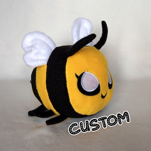 May include: A yellow and black plush bee with white wings. The bee has a smiling face with black eyes and eyelashes. The bee is sitting on a white surface with the word "CUSTOM" written in black on the surface.