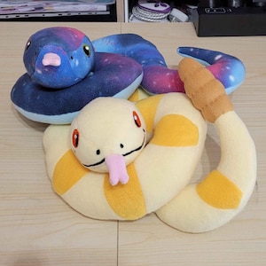 May include: Two plush snake toys. One is blue with a galaxy pattern, the other is cream-coloured with yellow stripes. Both have cartoonish features, including large eyes and pink tongues. The snakes are soft and cuddly.