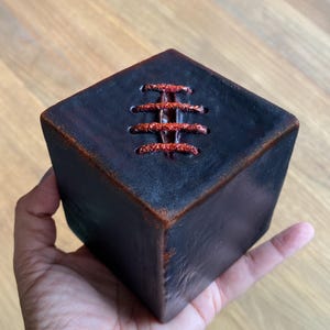 May include: A dark, square object with a stitched design on top, held in a hand. The object is primarily black with reddish-brown accents and stitching. The stitching is a bright red color.