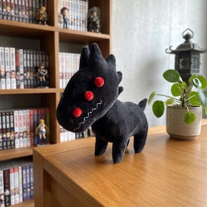 May include: A black plush creature with red accents, including three circular eyes and a zig-zag mouth. The plush toy has a stylized, cartoonish design and is displayed on a wooden surface. The creature has a tail and four legs.