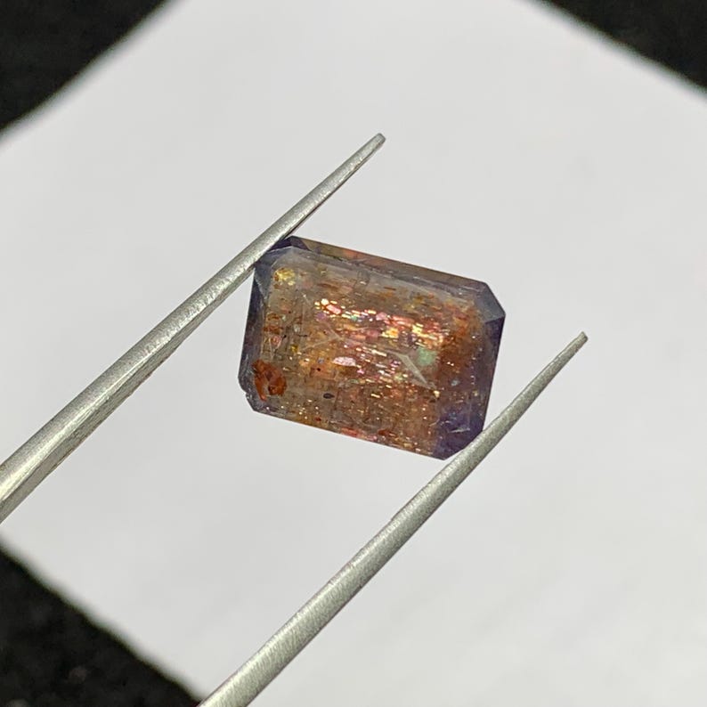 May include: A rectangular gemstone with a mix of colors, including purple, orange, and red, is held by tweezers. The gem has a faceted cut, and the colors shimmer. The gem is likely a sunstone or similar mineral.