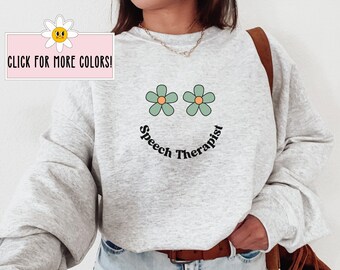 May include: Gray sweatshirt with a smiling face made of two green flowers and the text "Speech Therapist" underneath.
