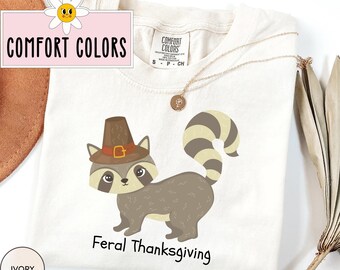 May include: A white t-shirt with a cartoon raccoon wearing a pilgrim hat and the text "Feral Thanksgiving". The shirt is from the brand "Comfort Colors" and is in the color ivory.