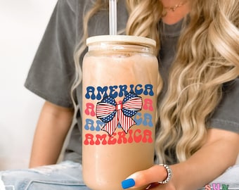 Può includere: Un bicchiere di vetro trasparente con un coperchio in bambù e una cannuccia. Il bicchiere ha una grafica di un fiocco rosso, bianco e blu con la parola "AMERICA" ripetuta in un font retrò.