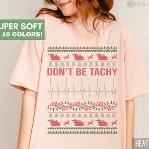 May include: A pink t-shirt with a Christmas-themed design featuring a red heart, a reindeer, and the text "DON'T BE TACHY" in red. The text is surrounded by a green and brown pattern.