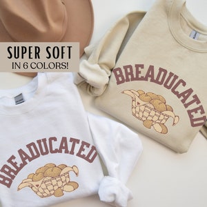 May include: Two sweatshirts with the text "BREADUCATED" in brown lettering. The sweatshirts are white and beige. The design features a basket of bread rolls.