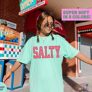 May include: A turquoise blue t-shirt with the word "SALTY" printed in pink letters. The shirt is super soft and available in 8 colors.