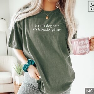 May include: Moss green t-shirt with the text "it's not dog hair it's labrador glitter". A pink mug with "Cool Mom Club" printed on it is held in the hand. The shirt is a casual style.