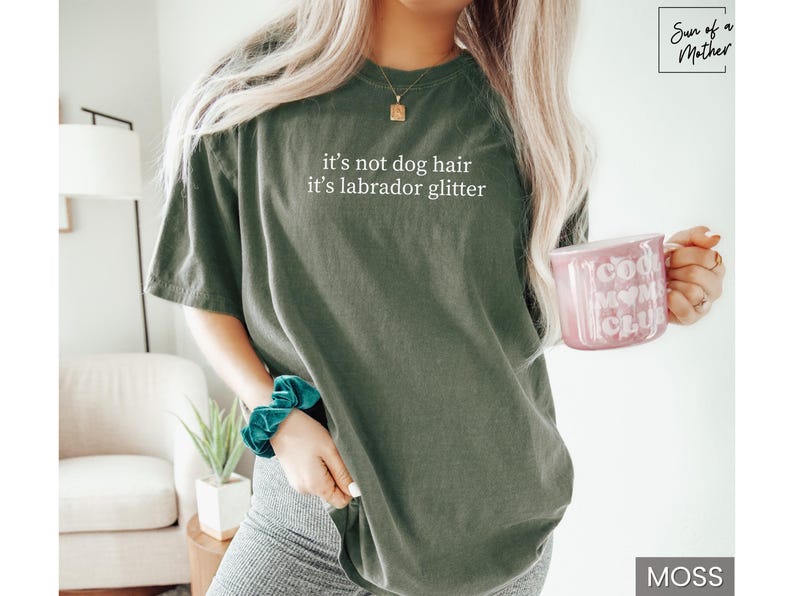 May include: Moss green t-shirt with the text "it's not dog hair it's labrador glitter". A pink mug with "Cool Mom Club" printed on it is held in the hand. The shirt is a casual style.