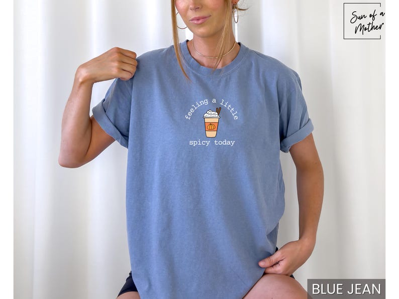 May include: Blue jean-colored t-shirt with rolled sleeves. The shirt features a graphic of a pumpkin spice latte with the text "feeling a little spicy today" in a whimsical font. The shirt is a comfortable, casual style.