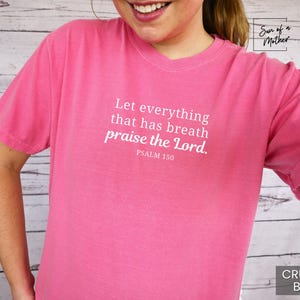 May include: A pink t-shirt with the text "Let everything that has breath praise the Lord. Psalm 150" in white. The shirt is a solid color and has short sleeves. The color is "Crunch-Berry".