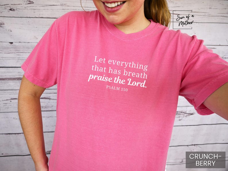 May include: A pink t-shirt with the text "Let everything that has breath praise the Lord. Psalm 150" in white. The shirt is a solid color and has short sleeves. The color is "Crunch-Berry".