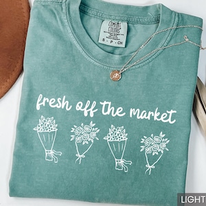 May include: Light green t-shirt with the text "fresh off the market" and four bouquets of flowers. A silver necklace with a small pendant is on the shirt. The shirt is labeled "Comfort Colors" and is size S-P-CH.