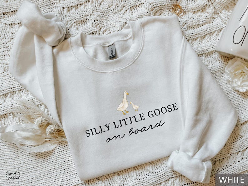 Peut inclure: Sweat-shirt blanc &agrave; col rond avec l'inscription "SILLY LITTLE GOOSE on board" en &eacute;criture noire. Le motif comprend une illustration &agrave; l'aquarelle d'une oie et d'un oison. Le sweat-shirt est de la marque Gildan.
