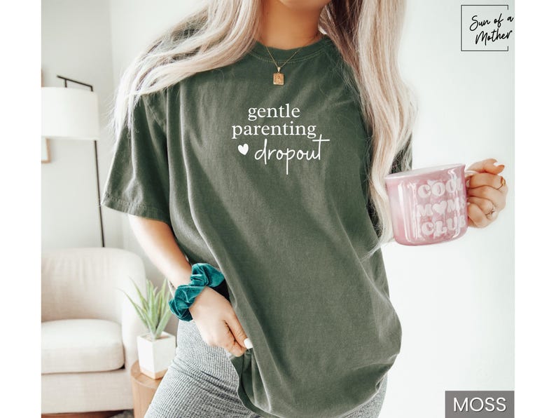 Puede incluir: Camiseta verde musgo con el texto blanco "gentle parenting dropout". Una taza rosa con "Cool Mom Club" se sostiene en la mano. Una scrunchie turquesa est&aacute; en la mu&ntilde;eca. La camiseta es de corte holgado.