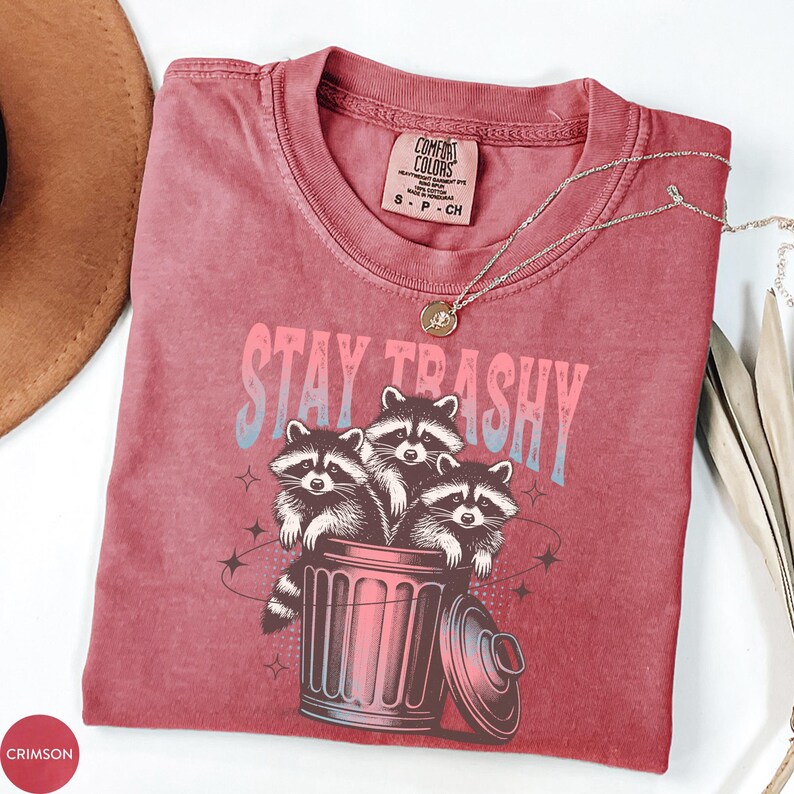 Op de afbeelding: Een rood T-shirt met een afbeelding van drie wasberen in een prullenbak. De tekst "Stay Trashy" staat boven de wasberen.