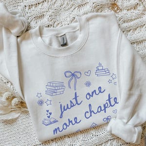 May include: White sweatshirt with the text "just one more chapter" in blue script. The design includes illustrations of books, hearts, stars, flowers, and a bow. The sweatshirt is a crew neck style.