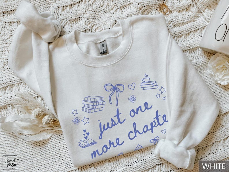 May include: White sweatshirt with the text "just one more chapter" in blue script. The design includes illustrations of books, hearts, stars, flowers, and a bow. The sweatshirt is a crew neck style.