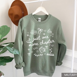 May include: Military green sweatshirt with the text "just one more chapter" in white. The design includes book illustrations, hearts, stars, and bows. A brown hat hangs on a white rack behind the sweatshirt.