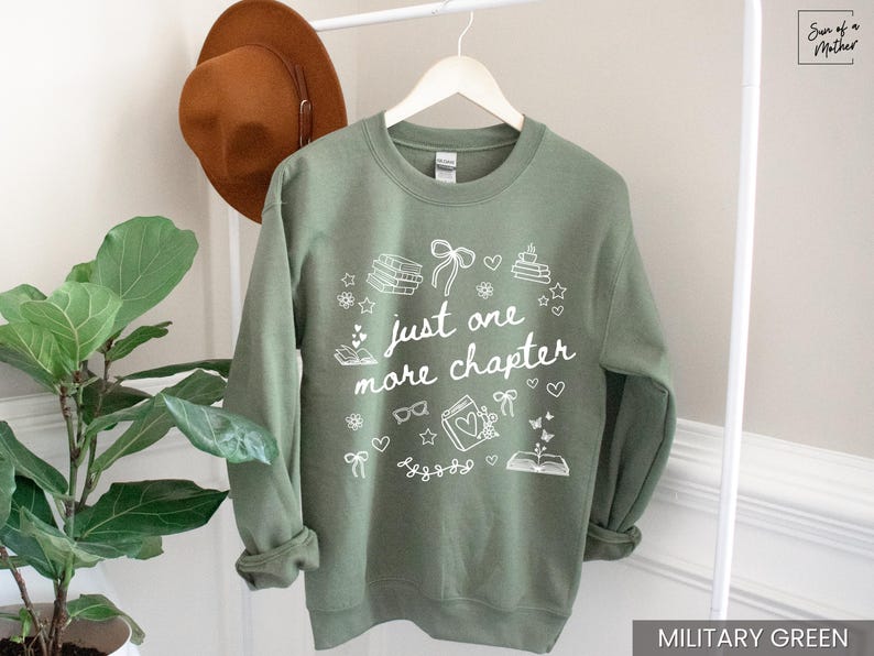 May include: Military green sweatshirt with the text "just one more chapter" in white. The design includes book illustrations, hearts, stars, and bows. A brown hat hangs on a white rack behind the sweatshirt.