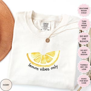 K&ouml;nnte beinhalten: Ein T-Shirt in Elfenbein mit einem Aquarell-Zitronenscheiben-Design und dem Text "lemon vibes only". Das Shirt hat einen Rundhalsausschnitt und kurze &Auml;rmel. Pflegehinweise sind auf der rechten Seite des Bildes aufgef&uuml;hrt.