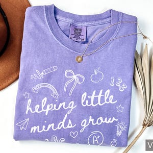 May include: Purple t-shirt with white text that says "helping little minds grow" and is surrounded by various school-themed illustrations.