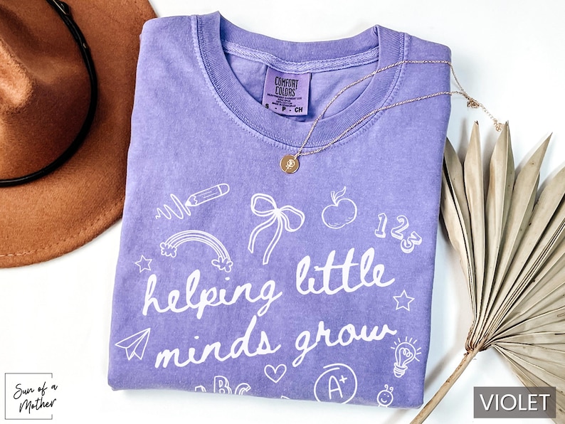 May include: Purple t-shirt with white text that says "helping little minds grow" and is surrounded by various school-themed illustrations.