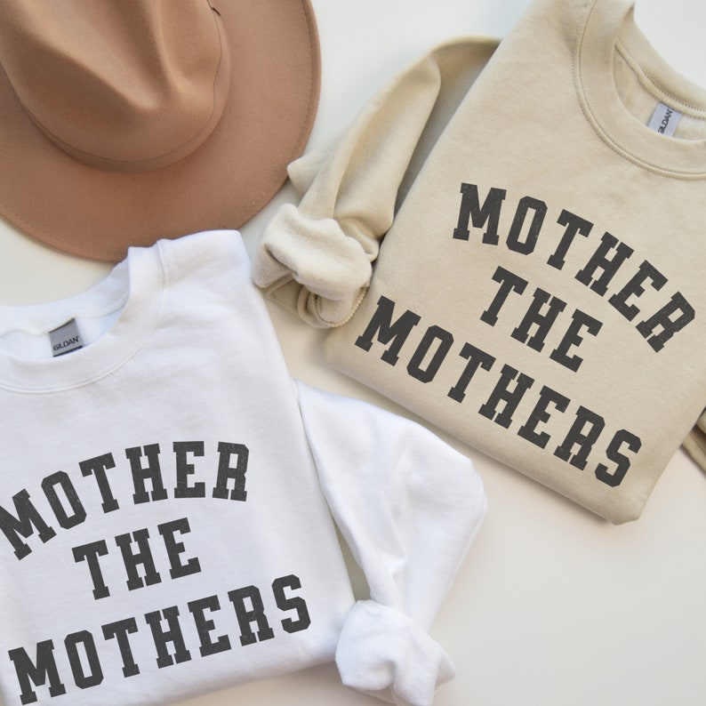 K&ouml;nnte beinhalten: Zwei Sweatshirts mit dem Text "Mother the Mothers" in schwarzer Schrift auf wei&szlig;em und beigefarbenem Hintergrund.