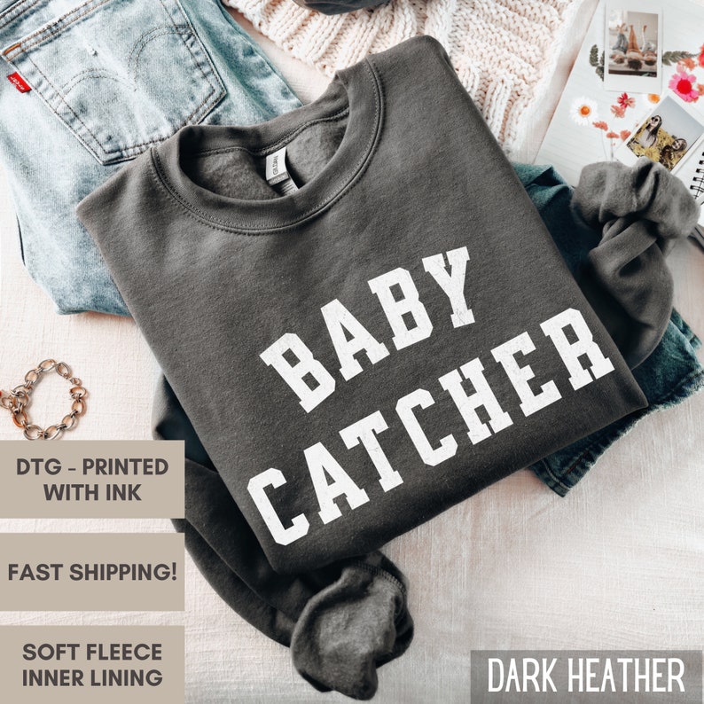 May include: Dark heather grey sweatshirt with the text "BABY CATCHER" printed in white.