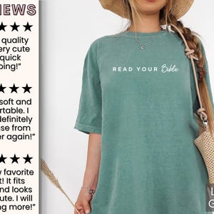 May include: A light green t-shirt with the text "READ YOUR Bible" in white. The t-shirt is made of a soft, comfortable fabric and has a relaxed fit. The model is wearing the t-shirt with a straw hat and a woven bag.