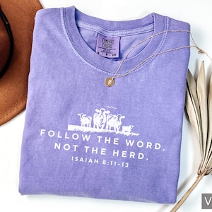Peut inclure: T-shirt violet avec un graphique blanc de moutons et le texte "FOLLOW THE WORD, NOT THE HERD. ISAIAH 8:11-13".