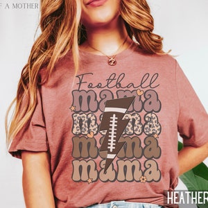 Puede incluir: Una camiseta de color malva brezo con el texto "Football Mama" en una fuente retro. El texto est&aacute; superpuesto con un estampado de leopardo marr&oacute;n y negro y un rayo marr&oacute;n con un bal&oacute;n de f&uacute;tbol en su interior.