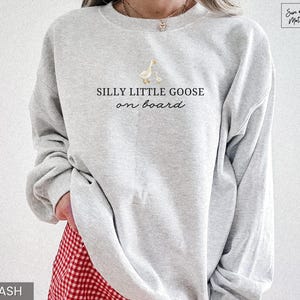 Peut inclure: Sweat-shirt gris clair avec l'inscription "SILLY LITTLE GOOSE on board" et un dessin d'une oie et d'un oison. Le sweat-shirt &agrave; manches longues a un col rond. Le bas du sweat-shirt est partiellement visible, et la couleur est "ASH".