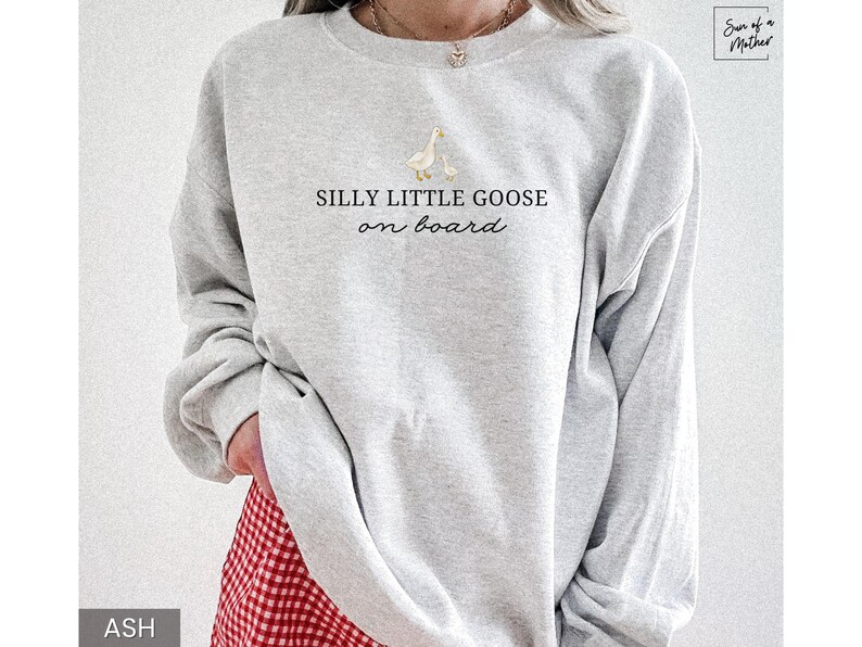 Peut inclure: Sweat-shirt gris clair avec l'inscription "SILLY LITTLE GOOSE on board" et un dessin d'une oie et d'un oison. Le sweat-shirt &agrave; manches longues a un col rond. Le bas du sweat-shirt est partiellement visible, et la couleur est "ASH".