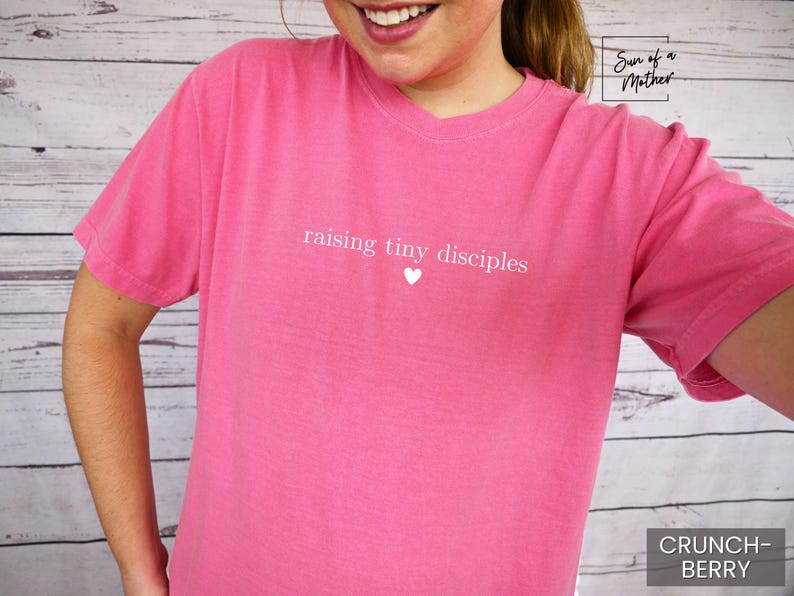 Op de afbeelding: Een roze T-shirt met korte mouwen met de witte tekst "raising tiny disciples" en een klein wit hartje. Het shirt is effen van kleur en heeft een ronde hals. De woorden "Crunch-Berry" staan in een wit kader rechtsonder.