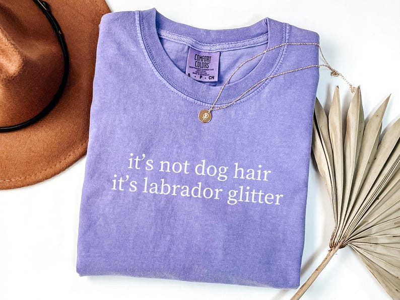 May include: A lavender Comfort Colors t-shirt with the text "it's not dog hair it's labrador glitter" in white. A gold necklace and a brown hat are also in the image. A dried palm leaf is in the background.