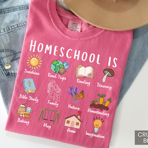 K&ouml;nnte beinhalten: Ein pinkfarbenes T-Shirt mit den Worten "Homeschool Is" und verschiedenen Illustrationen, darunter eine Sonne, ein Globus, ein Buch und ein Pilz. Das Shirt zeigt auch die W&ouml;rter "Sunshine", "Road Trips", "Reading" und "Discovery". Das Shirt liegt auf einer Jeans.