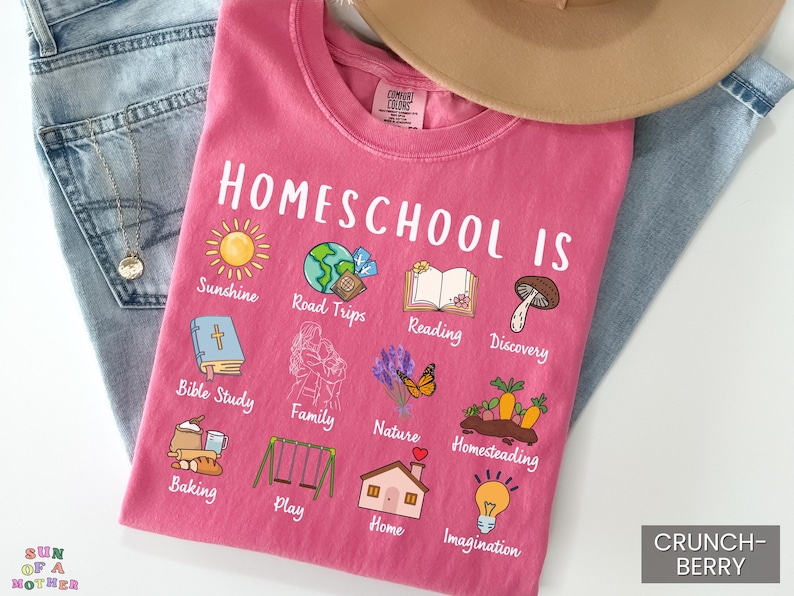 K&ouml;nnte beinhalten: Ein pinkfarbenes T-Shirt mit den Worten "Homeschool Is" und verschiedenen Illustrationen, darunter eine Sonne, ein Globus, ein Buch und ein Pilz. Das Shirt zeigt auch die W&ouml;rter "Sunshine", "Road Trips", "Reading" und "Discovery". Das Shirt liegt auf einer Jeans.