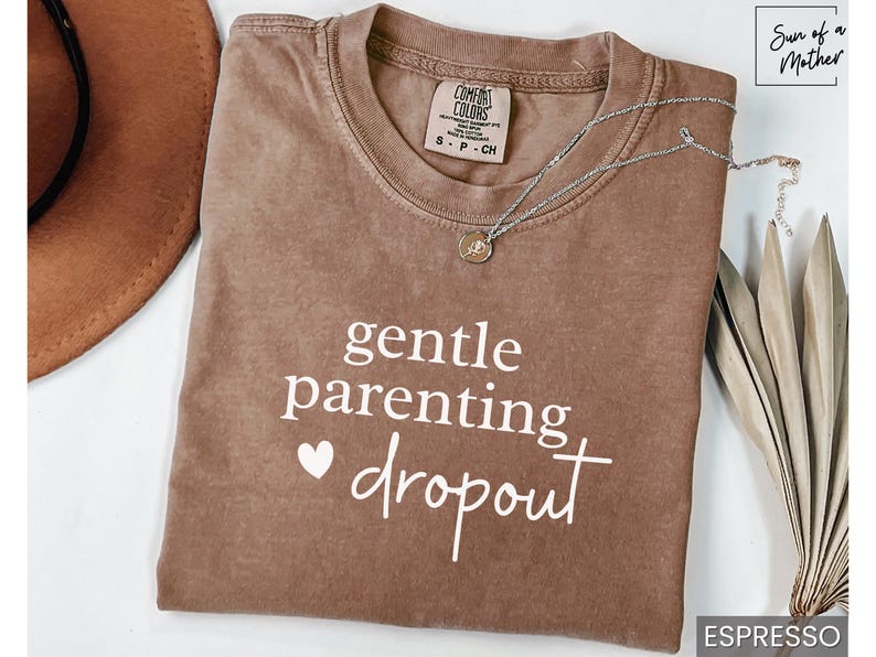 Puede incluir: Camiseta color espresso con la frase "gentle parenting dropout" en escritura blanca. Un collar plateado con un peque&ntilde;o colgante descansa sobre la camiseta. Un sombrero marr&oacute;n y una planta seca est&aacute;n en el fondo.
