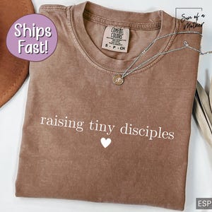 Op de afbeelding: Espresso-kleurig t-shirt met de witte tekst "raising tiny disciples", een klein wit hartje en een zilveren ketting. Het shirt is van het merk Comfort Colors, maat S-CH.