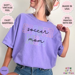 May include: Purple t-shirt with the text "soccer mom" in colorful outlined letters. The shirt is made of a soft, vintage feel fabric.