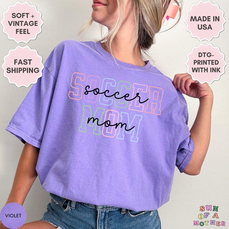 May include: Purple t-shirt with the text "soccer mom" in colorful outlined letters. The shirt is made of a soft, vintage feel fabric.