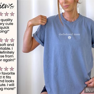 以下が含まれることがあります： ロールアップスリーブのブルーデニムカラーTシャツ。「dachshund mom」の文字と足跡のグラフィックがプリントされています。「Sun of a Mother」コレクションの一部です。