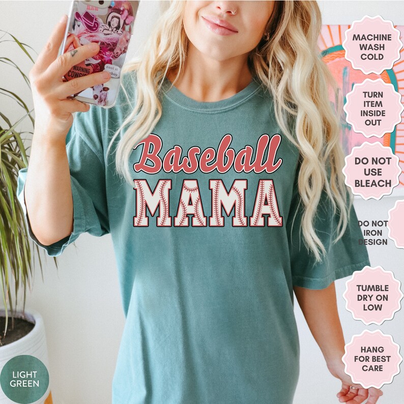 May include: A teal blue t-shirt with the text "Baseball Mama" in red and white. The text is surrounded by a baseball graphic. The shirt has care instructions printed on the right side of the image.